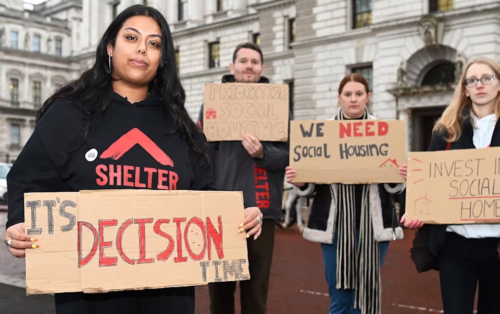 Group of housing campaigners outside a government building holding cardboard signs, including messages like It’s decision time, We need social housing, and Invest in social homes, during a protest organised by Shelter.